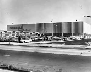 A black and white image of a Gimbels department store. A parking lot filled with vehicles and a road to enter the store's property is also depicted. 