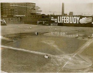 A black and white photograph of the baker bowl baseball field. The baseball diamond and field are wet with puddles around large sections of the field. A few players in uniform are running on the outfield, and to the stands on the left side of the image people are sitting. There are large brick buildings in the background and an advertising billboard for Lifebuoy soap. 