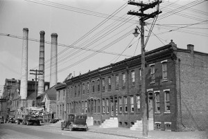 A black and white image of a  series of brick row homes. THere is a factory with three smoke stacks further down the street. A telephone pole, a car, and a truck carrying pieces of wood are in front of the houses. 
