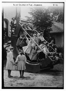 A black and white photograph of a group of children playing on a large piece of playground equipment. Two girls are standing in front of the equipment.