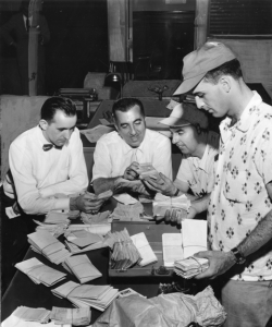 A black and white photograph of four men (three sitting and one on the right standing) looking over piles of receipts and money  on a table. 