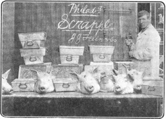 A black and white photograph of a man in white clothing standing to the right of a table filled with metal containers and hog heads. Written in chalk on a wooden surface behind the containers and heads are the words "Philadel Scrapple, J. J. Felin & Co.
