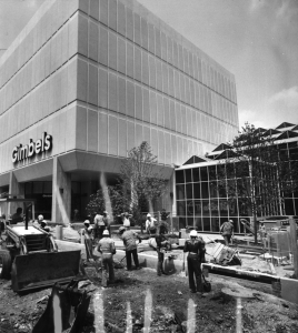 A black and white photograph of a construction crew outside of the glass and steel building, working on the sidewalk and road.