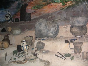 A color photograph of a variety of clay pots, jewelry, stone tools, metal tools, and other metal items are on an uneven brown landscape.  