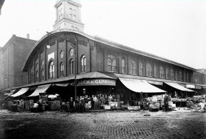 A black and white photograph of a building with a large tower at the front of the rectangular building. There is an overhang covering the sidewalk along the building, and the sidewalk is filled with people and products. 
