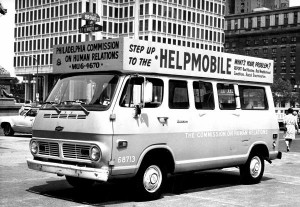 A black and white photograph of a van covered in signs and lettering parked on the street. the main sign on top of the van reads "Step up to the Helpmobile."