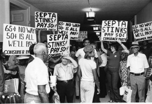 A black and white photograph of people standing in a hall way, holding protest signs. The group of people fills the hallway, and most of the people are wearing casual clothes. In the foreground of the image are some people with their backs turned to the camera.