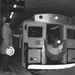 A black and white photograph of a man looking at the end of a subway car as it is leaving an underground subway terminal. The man is wearing a jacket, holding a hat, and standing by a large concrete pole that has a sign on it reading: