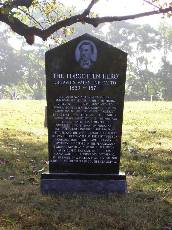 A color photograph of a black grave stone in the middle of a field. The stone has an engraving of a man at the top and a long epigraph covering most of the side facing the viewer. Some grass, leaves, and trees are visible.
