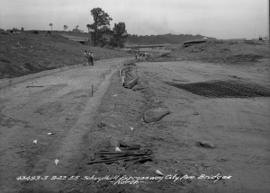 A photograph of some leveled dirt with markers and rope showing the edges of a road. Hills of dirt and people are in the background.