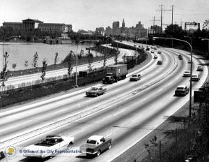 A black and white photograph of a highway with cars as it curves towards Philadelphia. Buildings, city hall, and the Philadelphia museum of art are in the background.