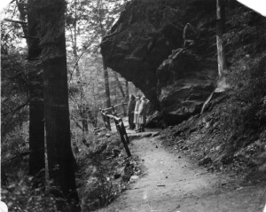A black and white photograph of three people standing on a trail below a rock ledge in the forest. 