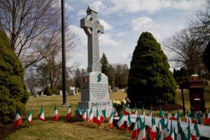 color photograph of a large celtic cross shaped headstone, surrounded by 57 Irish flags and a large plaque on the ground directly in front of it