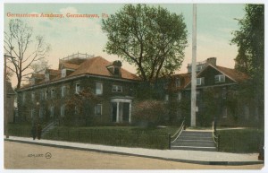 A color postcard of the corner of a city block, with some buildings and trees in the background. Some steps and a light pole are in the foreground and some people are walking on the sidewalk. 