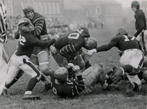 A black and white photograph of a group of high school kids playing football on a field. They are dressed in uniforms and small helmets. There is a small crowd in the background.