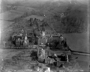 A black and white aerial photograph of a series of school buildings surrounded by fields and sections of trees. 