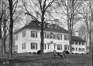 A black and white photograph of the front of a house, with trees and a cannon in the foreground.