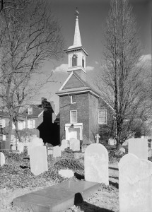 A black and white photograph of the front of a church, with a visible tower and graveyard stones in the foreground.