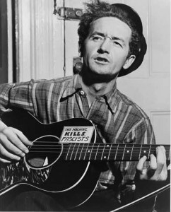 A black and white photograph of Woody Guthrie playing a guitar with the words "this machine kills fascists" written on it