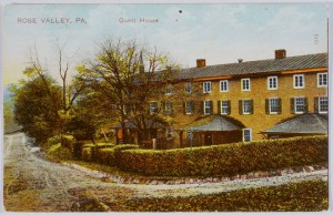 Scan of a postcard, in color. The valley rose guesthouse is photographed from an angle. The building is bright yellow with two rows of small white windows. The building is surrounded by a short hedge and obscured by a few trees. The road wraps around the building in the lower half of the postcard.