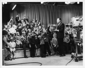 An image of Bill Webber speaking in front of cameras and an audience full of sitting and standing children. 