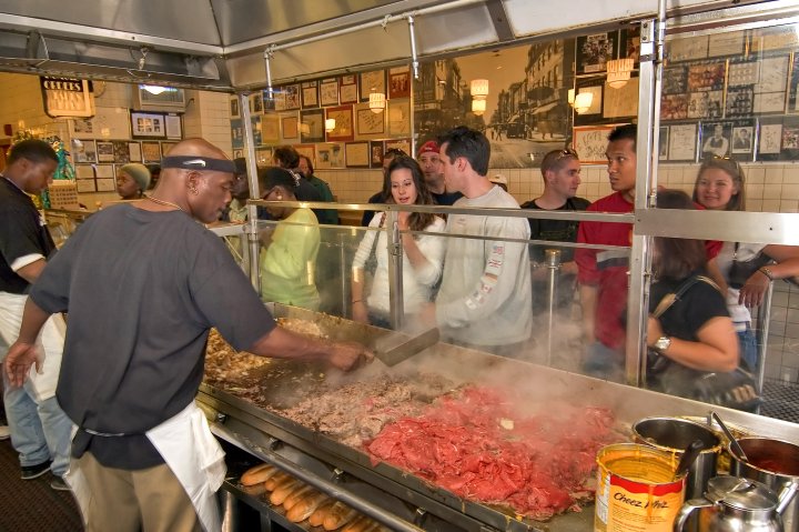 An photograph taken from behind the grill at Jim's Steaks. A chef is cooking cheese steaks while a group of people wait in line to purchase one.