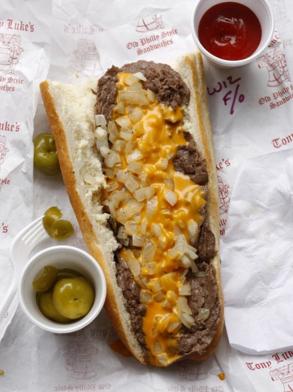 Photograph of a cheesesteak on a paper wrapper, with a cup of ketchup and a cup of peppers beside it. 