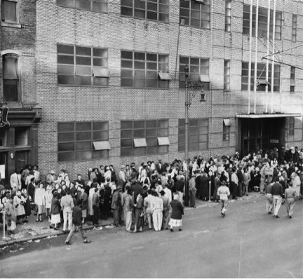 A large group of striking workers are standing outside one of the Campbell buildings; filling the sidewalk and blocking the doors of the building. 