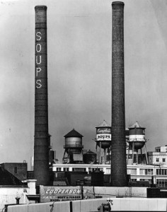 An image of two smoke stacks and three water towers of the Campbell's Soup Company building. The Water towers are painted with the Campbell's name and logo The Smoke stacks have the words 