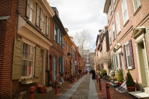 A color photograph of a row house-lined ally. The alley is paved with brick, and there are small concrete polls in front of each house. Each house has a variety of planters and small plants in front of the windows and next to the doors of each residence. The dominant construction material for these homes are brick, but wooden doors, shutters, and window frames are painted in various shapes of white, red, purple, and yellow. 