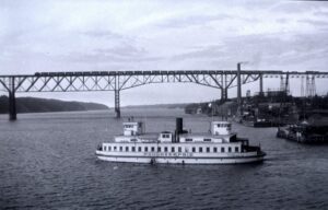 In the foreground, a ferry affixed with the name "Poughkeepsie" is traveling across a body of water with industrial imagery on the right and trees on the left. In the background is a train traveling across a bridge the spans the entire photograph.