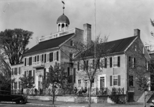 A large, three-sectioned Gregorian-styled building topped with an open cupola. An early twentieth-century car is parked in front.