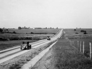 Two early twentieth-century cars drive in opposite directions along a skinny, paved road with farm land sprawled on each side of the road.