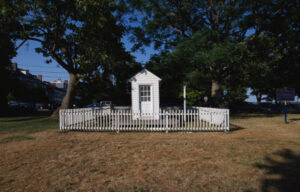 A small white shack surrounded by picket fencing. A door with a six-paned window is visible, which takes up most of the width of the shack.