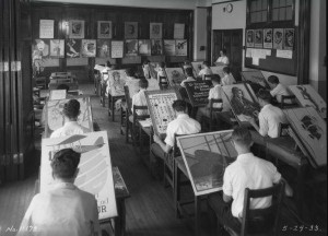 A photograph of a classroom with about eighteen students divided into four rows. Each student is sitting at their desk and they are all painting different images. A teacher is standing to the right of the students. A blackboard filled images is in the background of this image. 