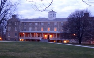 A color Photograph of the front of a three-story brick building in the evening. It is a long buildings with windows spaced evenly on the front of the building. There is a small tower at the top center of the building. Some trees and grass cover the area in front of the building. 