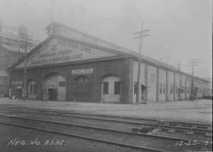 A black and white building of the Washington Avenue Immigration station, which was a long rectangular building with a center-peaked roof. The building's name is printed on the short side of the building, which is facing the front of the image, and there are some train tracks in front of the building. 