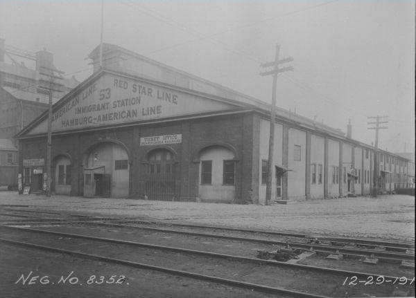 A black and white building of the Washington Avenue Immigration station, which was a long rectangular building with a center-peaked roof. The building's name is printed on the short side of the building, which is facing the front of the image, and there are some train tracks in front of the building.