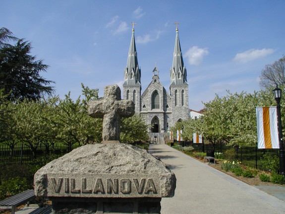 A color photograph of the front of St Thomas of Villanova church on a sunny day. There is a stone column with cross on top towards the foreground of the image, and the church is in the far background. Trees line the walkway up to the front of the church. 