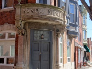 A color photograph of the front of the Banca D'Italia building. The Words "Banca D'Italia" are carved into the stone above the front door. This image also shows some of the brick siding of the bank, and some of the glass windows with stone designs above them. 