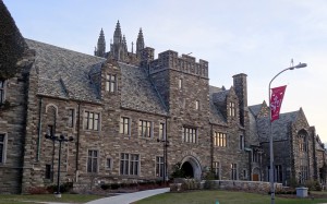 An color photograph of the front of a stone building. The stone is various shades of brown, sandstone, and grey. There is a walkway leading to the front door of the building. The building has a large tower in the back of the building, and spiral columns on the corners of the peaked roof. 