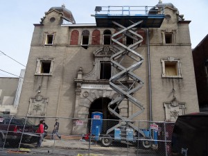 A color photograph of the front of a sand-colored stone Synagogue. There is a truck, a few people standing around, and an lift-device in front of the synagogue doing for some construction on the building. 