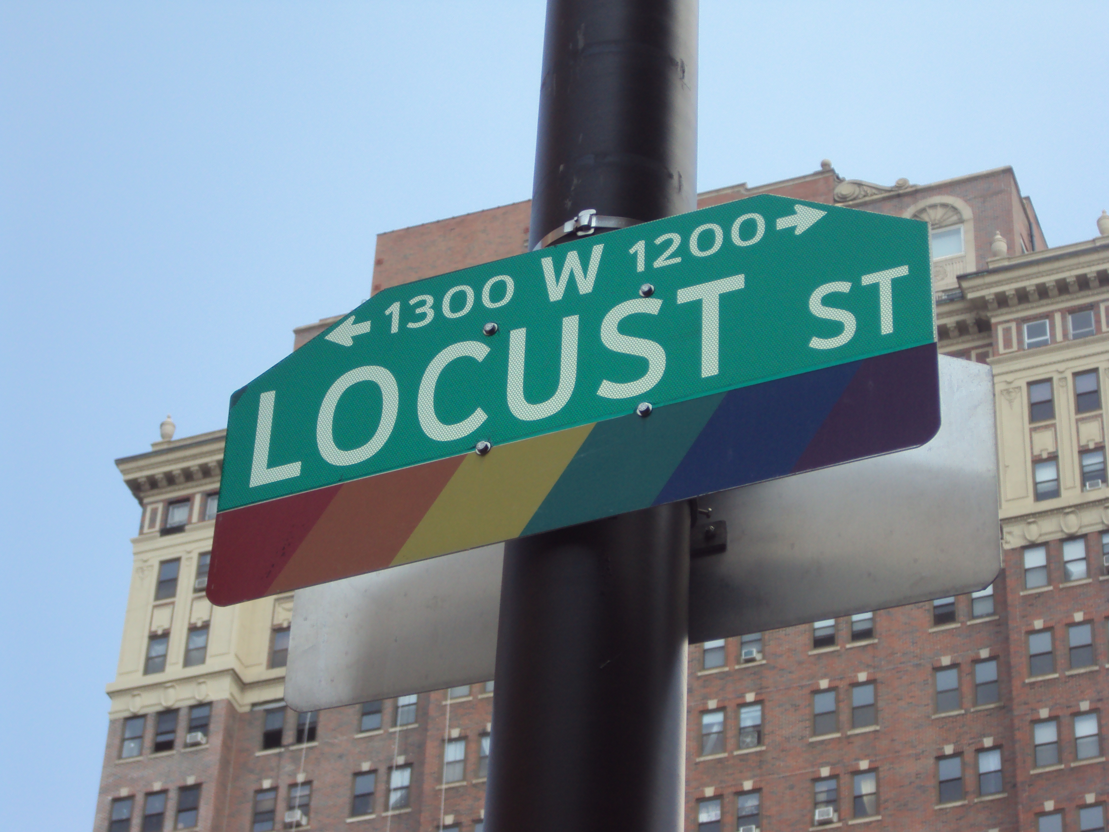 A color photograph of a street sign for Locust Street on a black pole. The green sign with white letters has an additional rainbow colored section on the bottom.