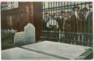 A color photograph of a group of people (many wearing hats) looking through a iron bar fence at the gravestones of Benjamin Franklin and his family. Some of the brick wall and other gravestones are visible on the left side of the image.