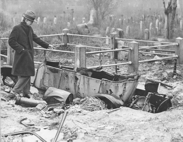 A black and white photograph of a man loking at an abandoned vehicle left in a graveyard. The grass is over gown, gravestones are knocked over, and the ground is uneven. There are also other metal parts surrounding the vehicle and scattered around through the background .