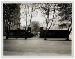 A black and white photograph of the front entrance to a cemetery. The brick walls and iron entrance are centered in the photograph. The foreground of the image shows the sidewalk and the paved street in front of the cemetery entrance. Trees are growing out of the curb of the foreground sidewalk and in the far background. 