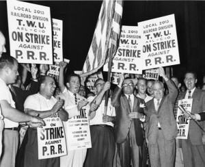 A black and white photograph of a crowd holding signs and banners displaying slogans for the strike. A man in the center of the image is holding an American flag.