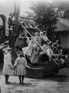 A black and white photograph of a group of children playing on a large piece of playground equipment. Two girls are standing in front of the equipment. 