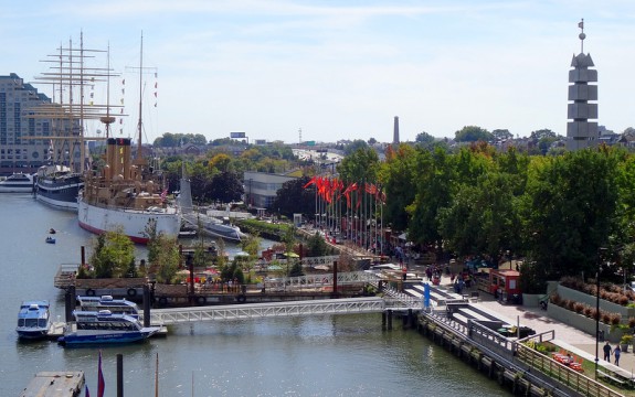 A color photograph of Penn's Landing, with ships on the water, a boardwalk with people walking on it, green trees and a steel obelisk above the treetops on the right side of the image. A older sailing ship is in the background of the image, along with more trees and buildings. 