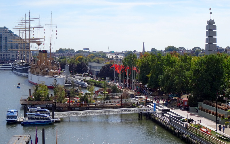 A color photograph of Penn's Landing, with ships on the water, a boardwalk with people walking on it, green trees and a steel obelisk above the treetops on the right side of the image. A older sailing ship is in the background of the image, along with more trees and buildings.